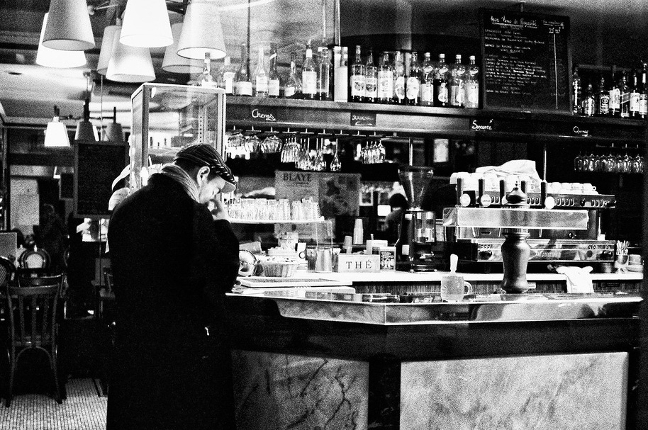 A person in a coat and cap stands at a coffee bar in a dimly lit café, savoring a petit noir amid bottles, glasses, and a coffee machine, with reflections shimmering in the glass behind the counter.