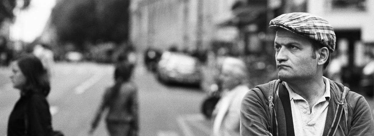 Black and white photo of a homme à la casquette in a plaid cap and jacket, standing on a city street with a serious expression; blurred pedestrians and cars move through the background.