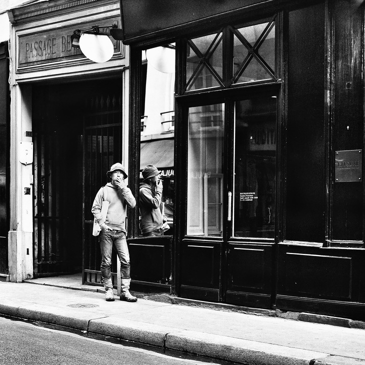A person in a hat stands on a quiet sidewalk by a shopfront, smoking a cigarette—perhaps recalling a passage à tabac. Their reflection appears in the black-and-white scene’s large window, while the empty street enhances the moody atmosphere.