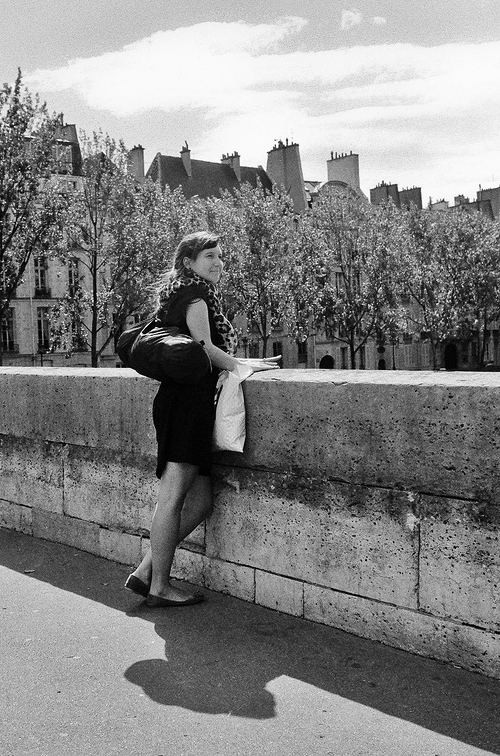 L'été c'est joli pour se parler d'amour A woman in a dress stands by a stone wall on a sunny été day, holding a bag and a paper, her gaze filled with amour as she looks toward trees and old buildings. Her shadow is softly cast on the ground.