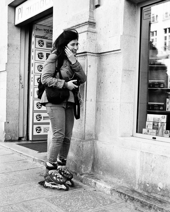 A patineuse souriant se tient sur un trottoir de la ville, tenant son visage d’une main tout en téléphonait. Elle porte un sac et se trouve près de l’entrée d’une pharmacie, avec des étagères et des enseignes en arrière-plan.