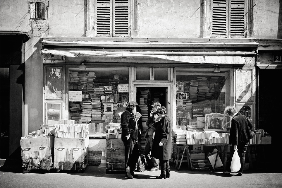 Black and white photo of La vieille echoppe, a vintage bookstore with stacks of books outside. Four people, dressed in coats and hats, browse the books on the sidewalk before the shop’s weathered facade and shuttered windows.