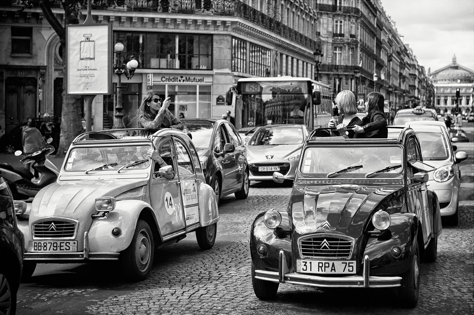 Two vintage Citroën 2CV cars with open tops cruise through an old school Paris traffic jam, surrounded by historic buildings. Two people in the cars snap photos and enjoy the vibrant city scene.