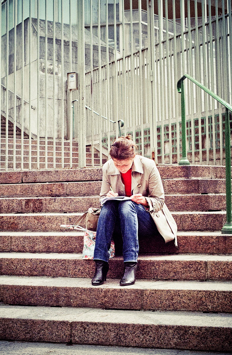 A woman in a light coat and jeans sits on outdoor stone marches, looking down and écriture in a notebook on her lap. She has a bag beside her, and metal railings are visible in the background.