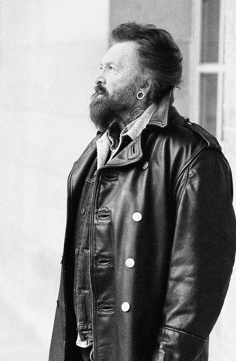 A bearded man with large ear gauges wearing a long black leather coat and layered denim clothing stands in profile, looking thoughtfully into the distance. The photo is black and white.