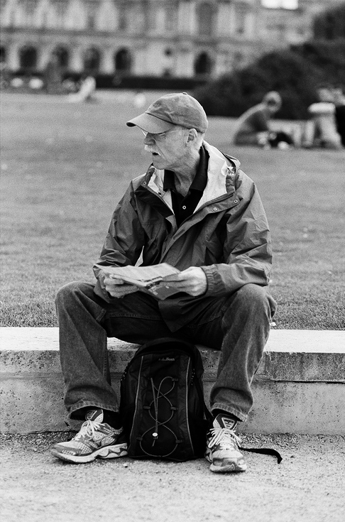 A man in a cap and jacket sits on a low stone wall outdoors, holding a map. A backpack rests at his feet and he looks to the side. The background shows grass, blurred buildings, and another person sitting on the lawn.