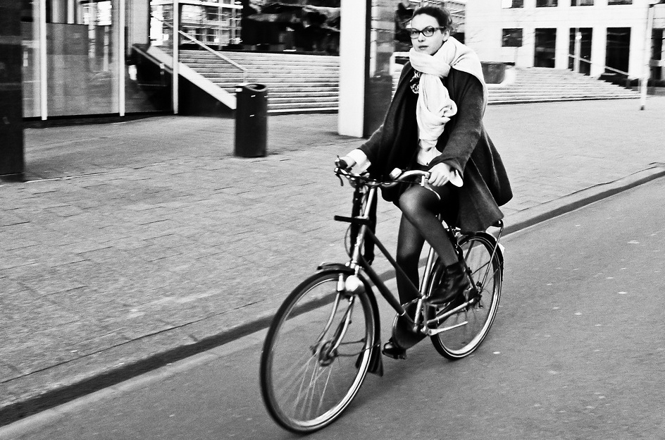 A person wearing glasses, a coat, and a large scarf rides a bicycle on a city street. Buildings and steps appear in the background. The image is black and white.