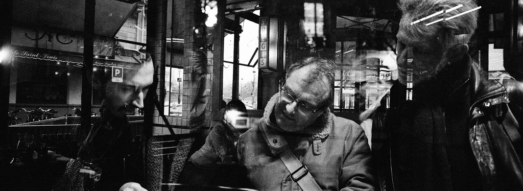 Des connaisseurs Three connaisseurs are seen through a reflective window, their faces partially obscured by glare and reflections. The scene appears candid and urban, giving a moody, layered effect in black and white.