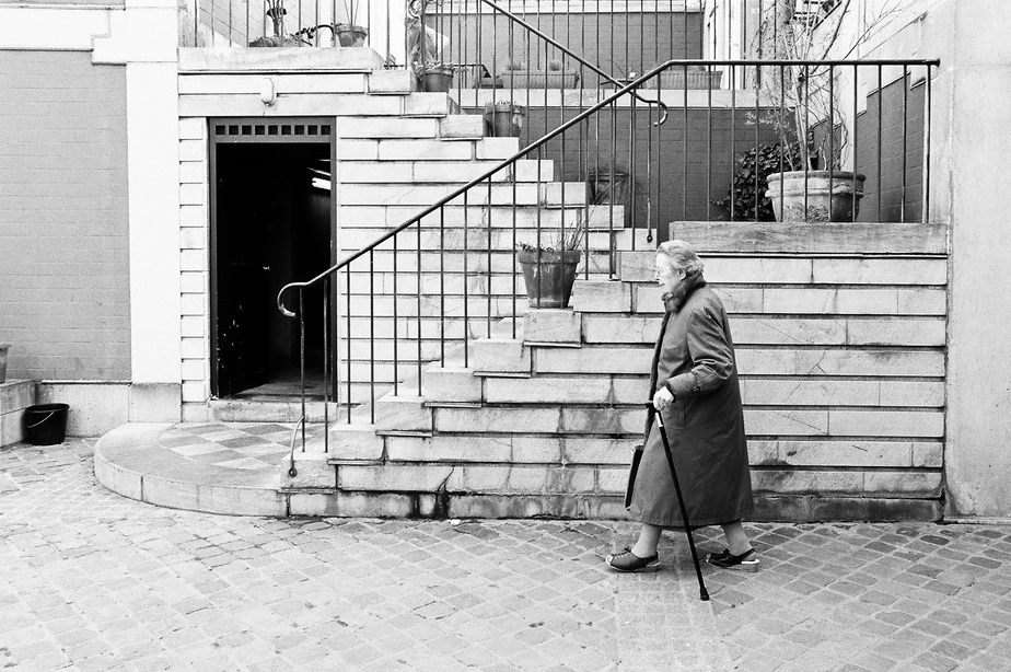 Black and white photo of Mme Clementin walking with a cane along a cobblestone path next to stairs with iron railings. Wearing a long coat, she appears to be outdoors, possibly in an urban setting with brick and stone buildings.