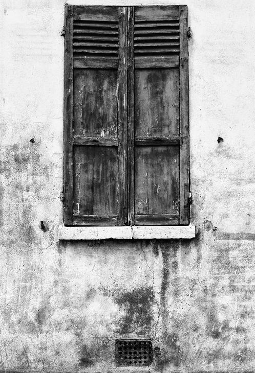 A black-and-white photo of an old, weathered wooden window with volets clos on a rough, stained concrete wall. A small vent is visible near the bottom of the wall.