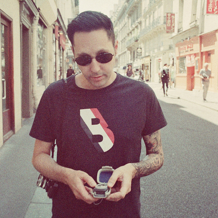 A man with dark hair and sunglasses stands on a sunny city street, looking down at an open ring box in his hands. He wears a black T-shirt with a bold graphic design and has a tattoo on his right arm.