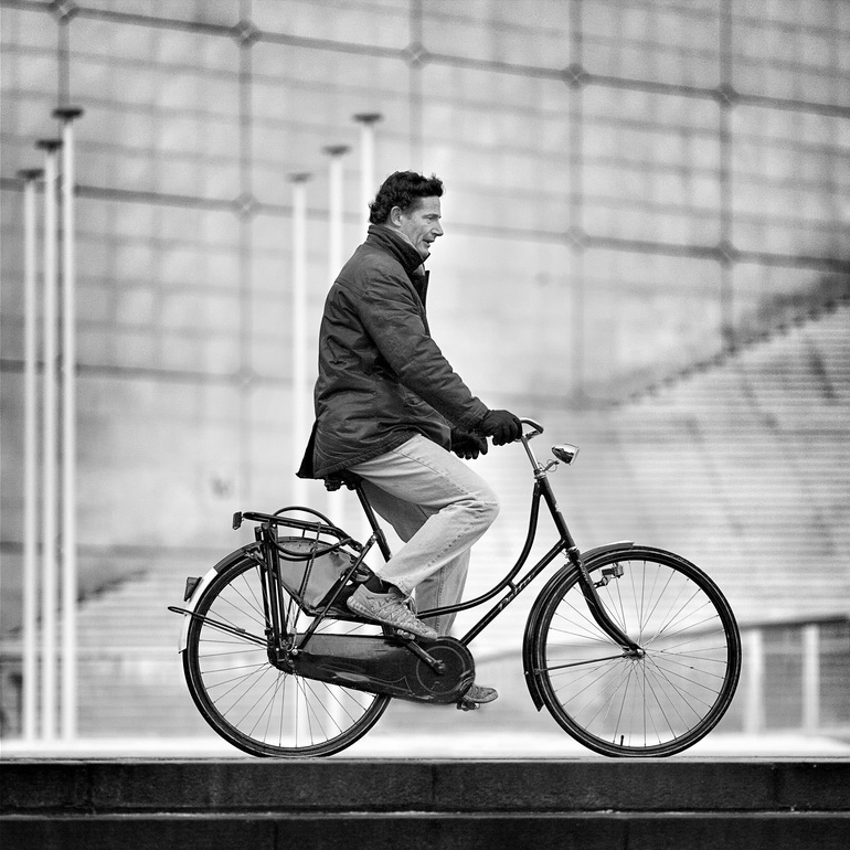A man wearing a jacket and gloves rides a bicycle in an urban setting, passing a geometric patterned wall, flagpoles, and stairs. Captured in black and white, this evocative scene is photographed by Paul Michalon.