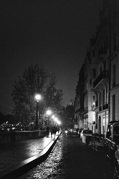 A cobblestone street at nuit, dimly lit by street lamps on île Saint-Louis. Silhouettes of people walk along the sidewalk beside parked cars and tall buildings; trees are visible on the left. The scene has a moody, atmospheric feel.