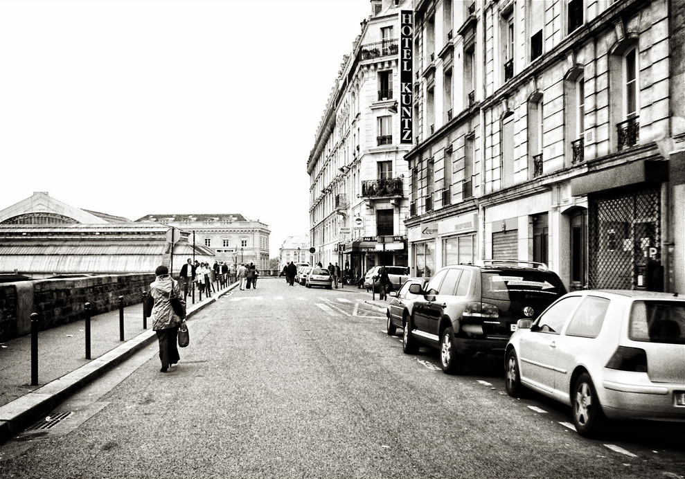 Black-and-white photo of Rue d'Alsace, with parked cars on the right, pedestrians on the left, and tall buildings including one featuring a prominent "HOTEL KUNTZ" sign in the background.
