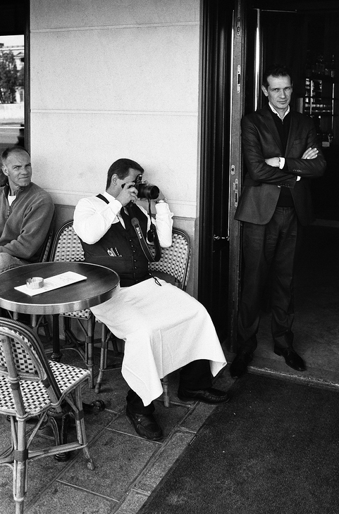 A black-and-white photo shows a garçon photographe sitting at a cafe table, looking through binoculars. Another man sits nearby, while a third stands by the doorway with arms crossed, watching the scene unfold.