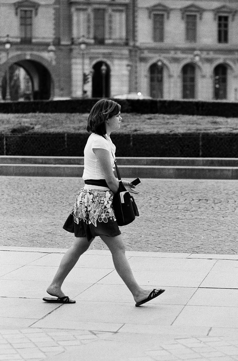 Delphine Mazeau, wearing a white shirt, patterned skirt, and flip-flops, walks on a paved street in front of a historic building with arched windows and ornate architecture. She carries a handbag and looks ahead.