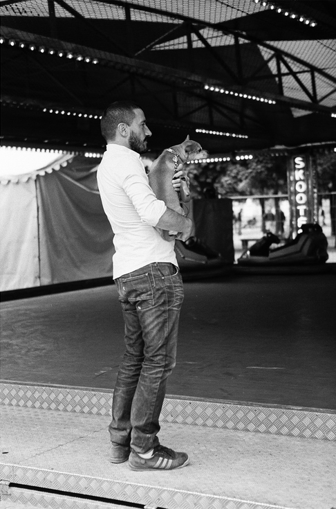 A man in jeans and a white shirt stands on the edge of an empty bumper car ride, holding a small dog in his arms. Captured in black and white, this striking scene by Cédric Ledoux highlights quiet moments amid stillness.