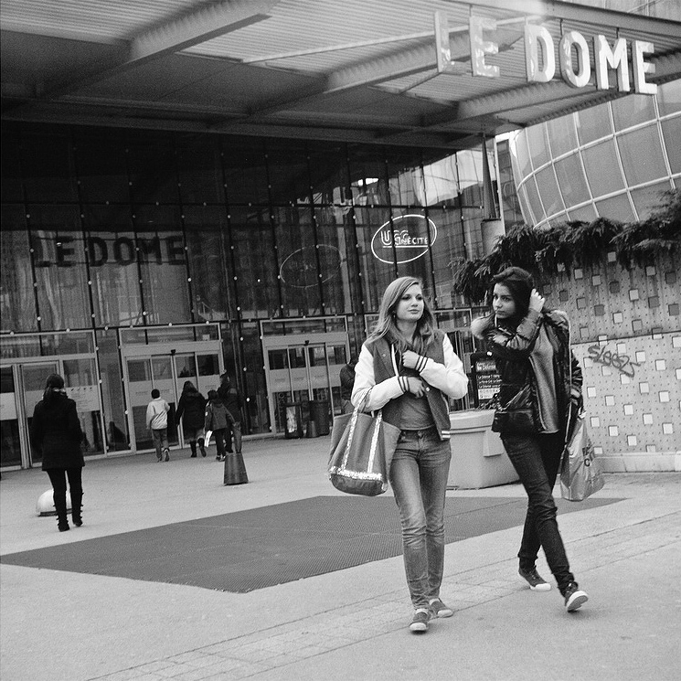 Two young women walk and talk outside a glass-fronted building with a sign reading "LE DOME." Other people are in the background near the entrance. The scene appears to be urban and lively.