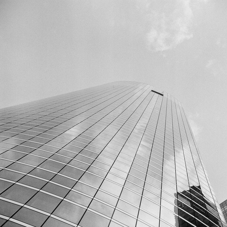 A black and white photo of a tall glass skyscraper seen from below, with reflections of the sky and clouds on its windows. In the lower right corner, Au pied de la tour, another building's silhouette is visible.