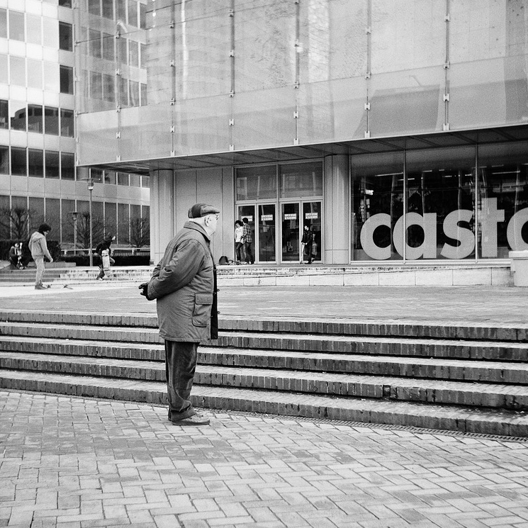 An elderly man, Pierre Lerond, stands at the bottom of a set of stairs, hands behind his back, looking toward a modern building with large glass windows. Several people walk near the entrance in the background.