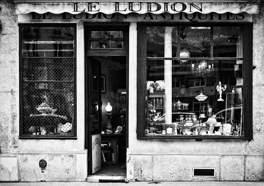 A black and white photo of a vintage storefront named "Le Ludion Antiquités." The display window shows various antiques including lamps, vases, and other decorative items. The door is slightly ajar, inviting people to explore the unique collection inside Le Ludion.
