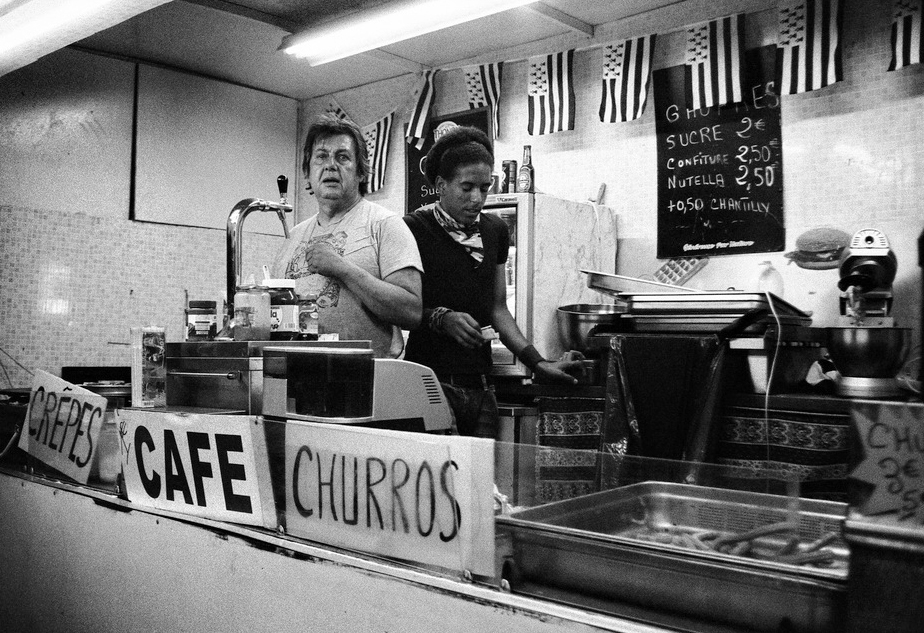 Two vendors stand behind the counter of a busy baraque à churros, selling crêpes, café, and churros. Handwritten signs and kitchen equipment add charm to the bustling scene, captured in black and white.