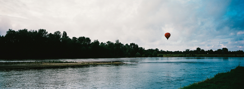 Un ballon sur la Loire A serene riverside landscape with a hot air balloon floating near the horizon. The ballon sur la Loire is adorned with a pattern resembling a ladybug. The river is calm, and lush green trees line the riverbank under a partly cloudy sky.