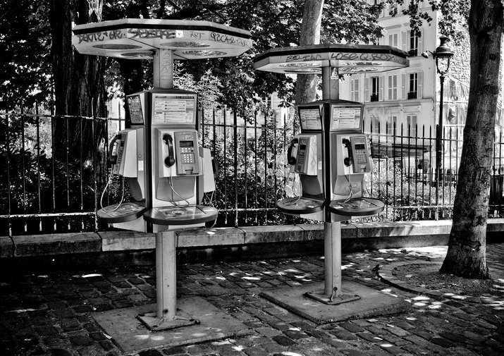 A black and white photo of Les deux cabines, two outdoor public payphones under large canopies. The payphones stand side by side on a cobblestone sidewalk, with a metal fence and trees in the background. Graffiti is visible on the canopies and nearby surfaces.