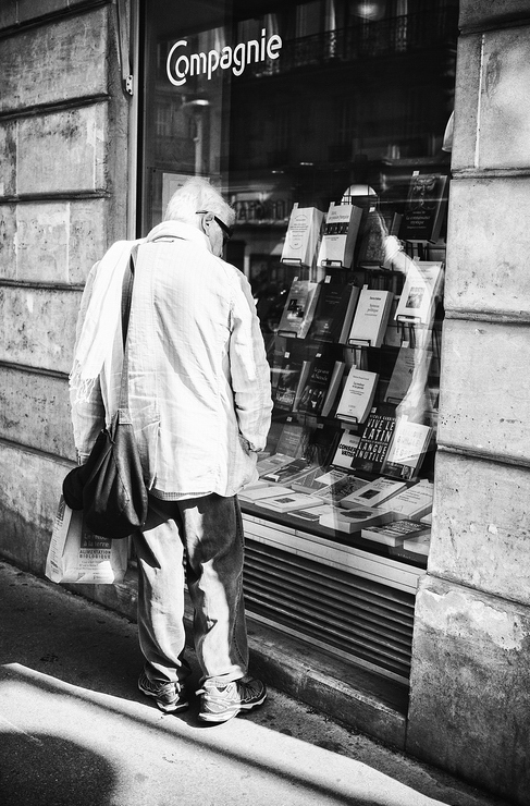 La bonne lecture A black and white image shows an elderly person standing on a sidewalk, looking into a bookstore window filled with various books. Wearing a light-colored jacket and carrying a bag over their shoulder, they gaze intently at the display. The store sign above the window reads "La bonne lecture - Compagnie.