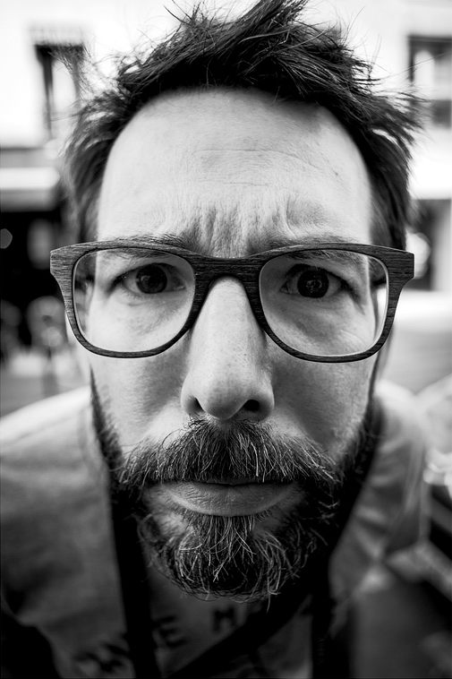 Intrigué Close-up, black-and-white photo of a man with glasses and a beard, leaning towards the camera with an intrigué and curious expression. The background is slightly blurred, highlighting the man's face and his inquisitive look.