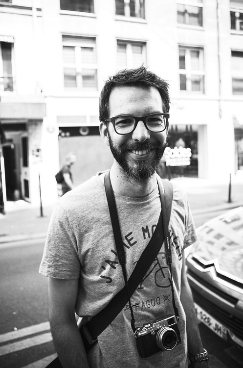 Xavier en mode photographe de rue Black and white photo of a smiling man with a beard and glasses, wearing a t-shirt and a camera slung around his neck. Photographe Xavier stands on a city rue with buildings and parked cars in the background.