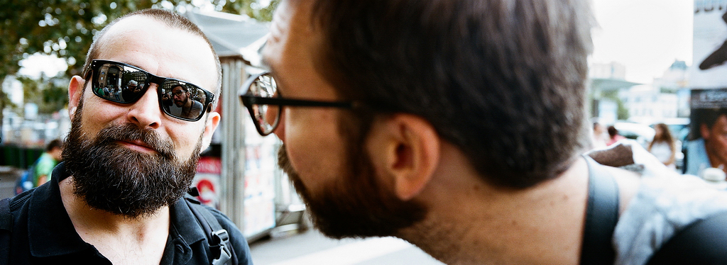 Oh on se voit dedans Two men with beards are seen in the image. One man, wearing dark sunglasses, looks off to the side while smiling slightly. The other man, whose face is not fully visible, leans in, appearing to be speaking or looking closely at the first man—"Oh on se voit dedans," one might imagine him whispering.