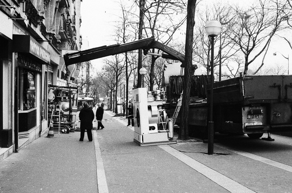 A black and white image captures a film set on a city sidewalk. Equipment and lighting are positioned near a building advertising "Grand Lessiveuse Pressing." Two people stand near the setup, while a crane extends over the sidewalk from a truck parked for delivery. Leafless trees line the street.