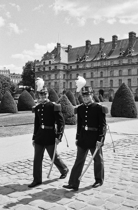 Two uniformed guards in formal attire, complete with feathered hats and sashes, walk side-by-side on a cobblestone path. They could be mistaken for Les frères Dessertine, standing before a historic, large building with manicured bushes and trees in the background. The scene is captured in black and white.