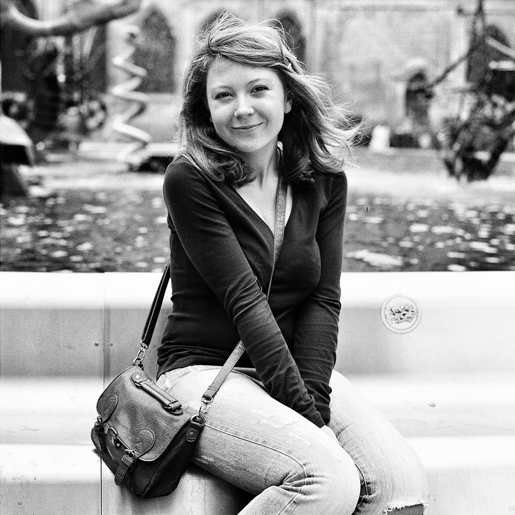 La soeur de GitFizz, with shoulder-length hair, smiles at the camera while sitting on a ledge in front of a fountain. She wears a dark long-sleeve top, jeans, and carries a crossbody purse. The blurred outdoor scene suggests an urban setting. The image is beautifully captured in black and white.