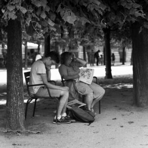 Two people sit beneath a tree, each reading—one a book, the other a newspaper. The seated man rests his head on his hand while the woman holds the newspaper open. A backpack lies on the ground between them. Sunlight filters through the leaves above.