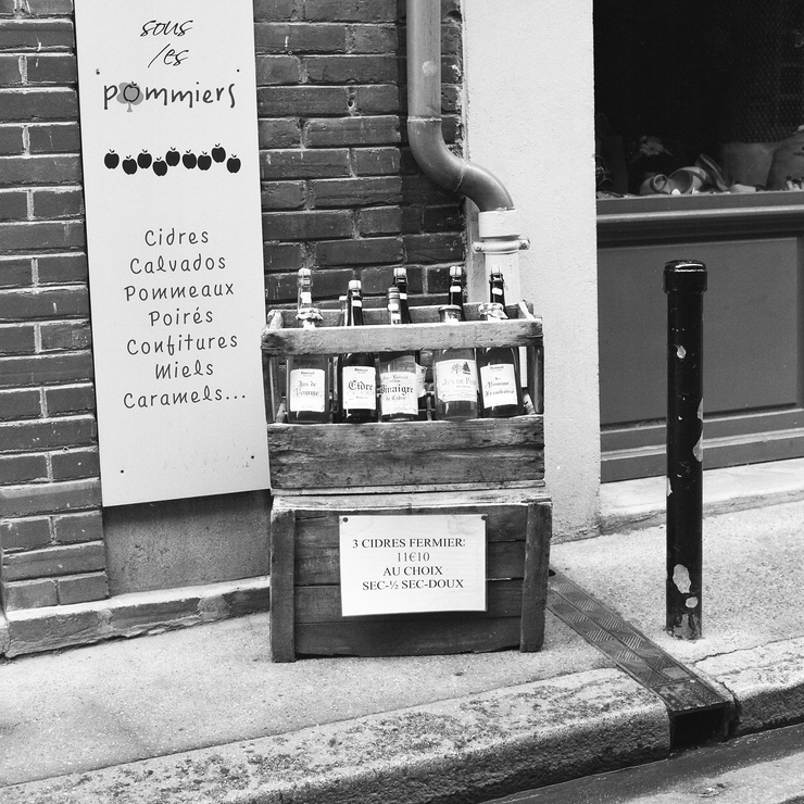A black and white photo of a street scene features a wooden crate filled with labeled bottles placed on the ground next to a building. A sign above the crate offers cider varieties, along with other products like honey and jams. Sans Titre is written inconspicuously on a wall near some visible pipes and bricks.