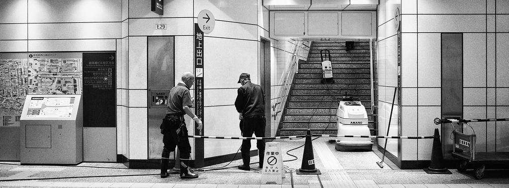 Black and white photo of two workers in a Tokyo métro station hallway. They are near a sign reading "Exit" with an arrow. A cleaning machine sits at the base of a staircase blocked off by tape and a sign with Japanese text.