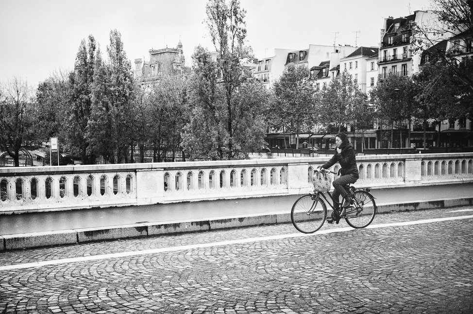 J'ai froid, je remonte mon echarpe A black and white photograph shows a person riding a bicycle on a cobblestone path across a bridge lined with ornate railings. Trees and buildings stand in the background, creating an urban landscape. The cyclist, wrapped in an écharpe against the froid, carries a basket on the front of the bike.