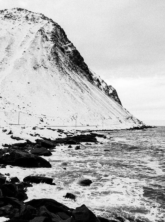 Myrland – Lofoten Islands – Norway A black and white image of a rugged, snow-covered cliff in the Lofoten Islands descending into a rocky coastline. The ocean waves crash against the rocks, with a narrow road winding along the base of the cliff. The sky is overcast, adding to the stark, dramatic atmosphere of Myrland, Norway.