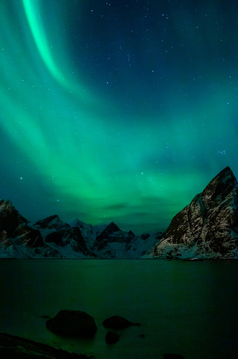 Aurore boréale / Hamnoy / Lofoten / Norvège A striking Aurore boréale lights up the night sky with vibrant green hues over the serene, snow-covered peaks of Lofoten. The scene reflects off a calm body of water in Hamnoy, creating a mesmerizing and tranquil atmosphere.