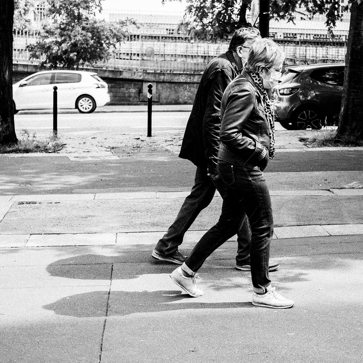 A black-and-white photo shows two individuals walking along a sidewalk. A woman with a scarf and leather jacket leads, while a man follows closely behind. Trees and parked cars are visible in the background, their shadows cast on the pavement—a quiet moment of Paris confiné.