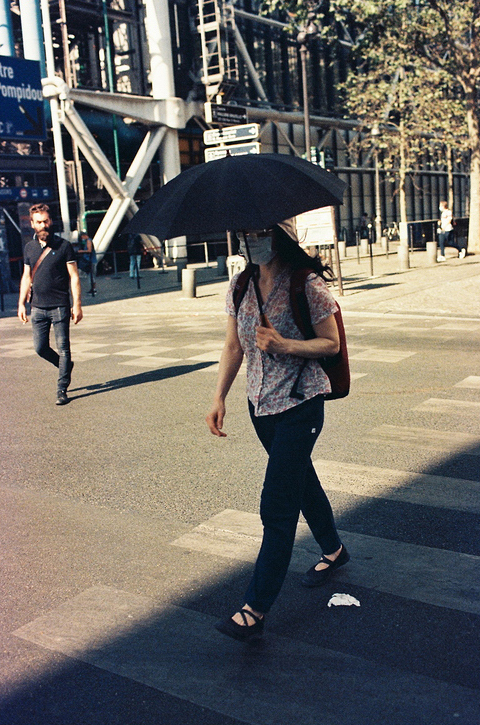 A person holding an ombrelle walks across a street intersection on a sunny day. They are dressed in a floral shirt, jeans, and sandals, with a red bag over their shoulder. Another person walks in the background, and a large building is visible behind them.