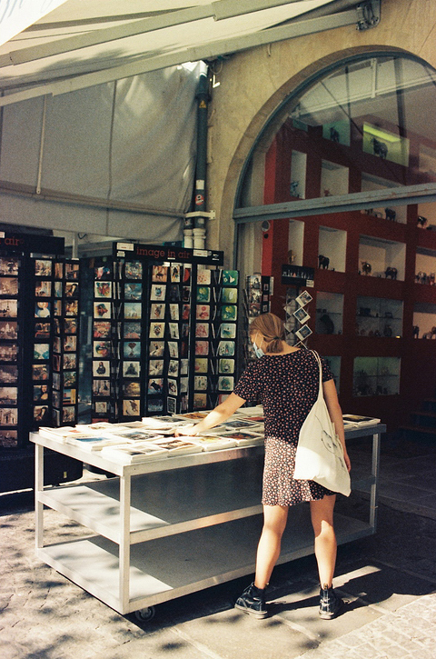 A woman wearing a patterned dress and a white tote bag, possibly by designer Lucie Dolce, browses through postcards on a metal table under a canopy. Behind her, postcard displays and a red wall with various items can be seen. The scene is lit by natural sunlight.
