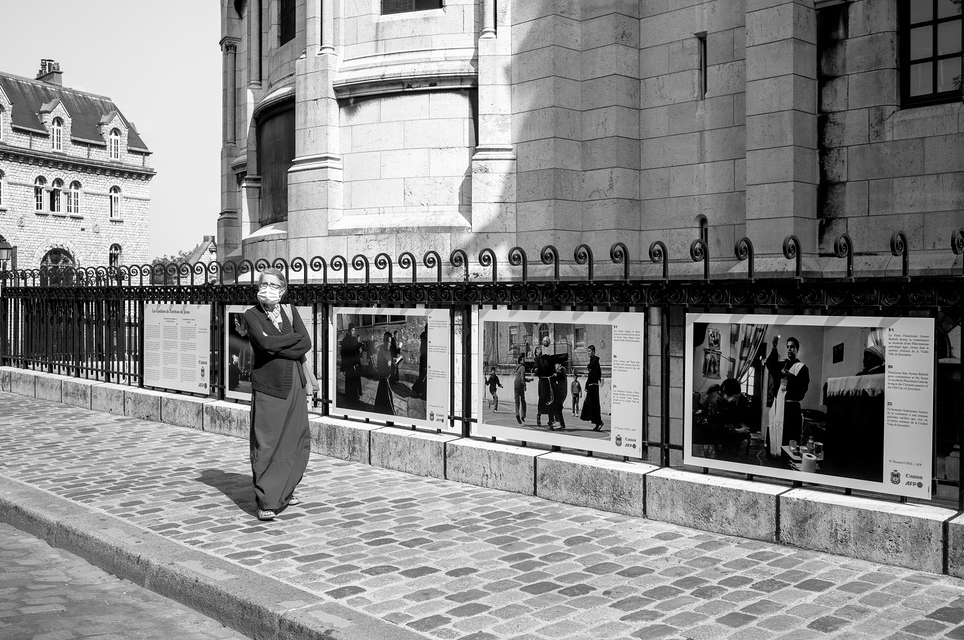 Montmartre A person wearing a long dress walks along a cobblestone street in the charming neighborhood of Montmartre next to an iron fence adorned with historical photographs. The background features a stone building with arched windows and architectural details, capturing the timeless essence of Paris in black and white.