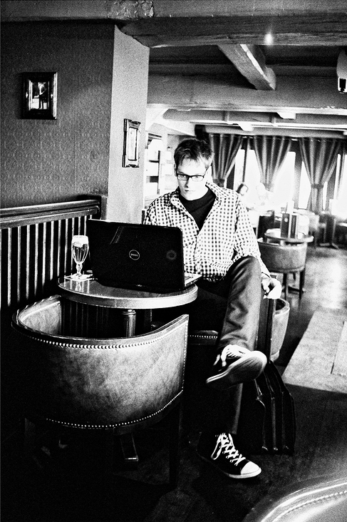 A person wearing glasses and a checkered shirt is sitting at a small round table in a dimly lit room, working on a laptop. A glass of wine is on the table beside them. The espace de travail has leather chairs, wooden beams, and curtains. The vibe is cozy and vintage.