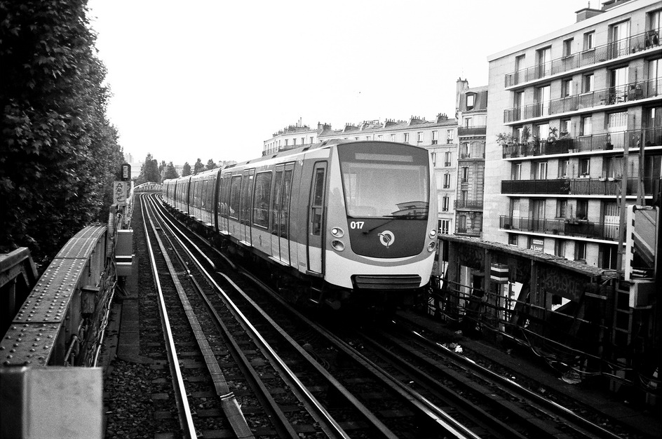 A black-and-white photo of a modern train on Ligne 2 traveling on an elevated track through an urban area. The train, composed of multiple cars, is surrounded by buildings and trees, capturing the front side as it moves forward.