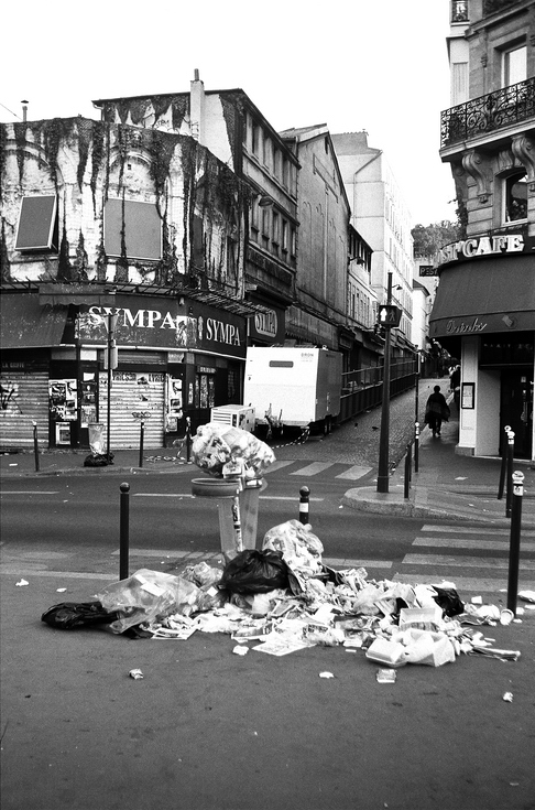 Black and white photo of a street scene showing an overflowing trash bin with détritus scattered around it. In the background, there is a shuttered building with a sign "Sympa" and a narrow street leading uphill. A few people are walking in the distance.