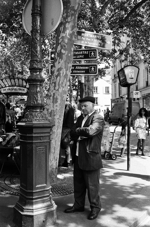A man in a striped suit and beret, resembling Jean de Montmartre, stands with arms crossed under a tree near a lamppost in Montmartre, Paris. Directional signs and people walking are visible in the background, along with a metro entrance. The scene is captured in black and white.
