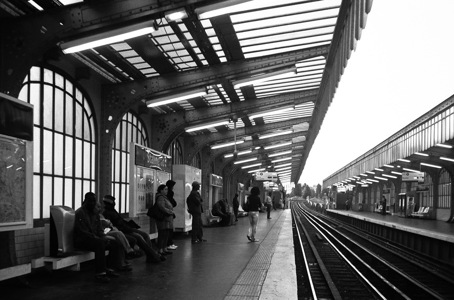 Black and white photo of a train station platform with several waiting passengers. The platform has an overhang with glass and metal detailing. Parallel tracks, part of Ligne 2, run to the right, and another platform is visible across the tracks.